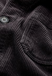 Schwarzer Cord-Stoff mit vertikalen Rippungen, versehen mit einem runden schwarzen Knopf und einer aufgesetzten Tasche. Die Textur ist weich und plüschig.