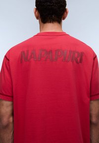 Κόκκινο βαμβακερό T-shirt με τη λέξη "NAPAPIURI" σε στιλιζαρισμένα, κεντημένα γράμματα στην πλάτη. Κοντά μανίκια, στρογγυλή λαιμόκοψη.