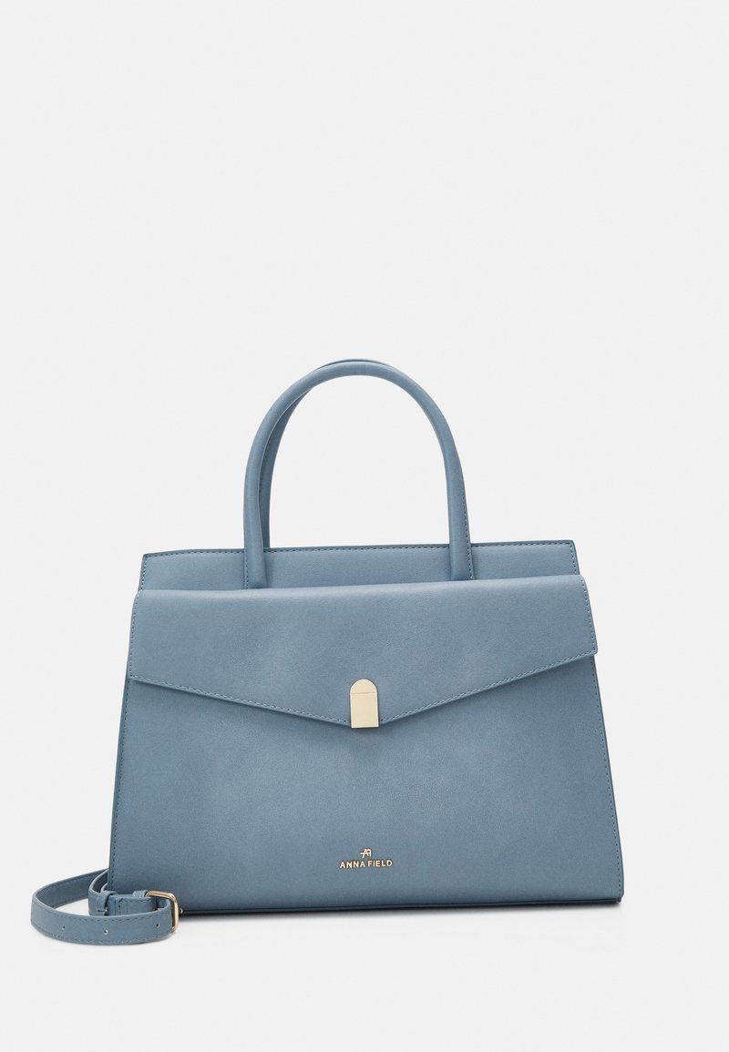 Sininen tekonahkainen käsilaukku, jossa on ryhdikäs muotoilu, yläkahvat, magneettisuljin ja irrotettava olkahihna. Pohjassa on logot yksityiskohta.