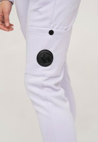 Pantalons violet clair avec une poche latérale, dotés d'un détail en patch noir rond. Fabriqués en tissu lisse avec une texturation subtile.