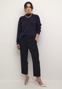Marineblauwe gebreide trui met lange mouwen, gecombineerd met zwarte op maat gemaakte broek en zilveren puntige hakken. Chunky metalen ketting en oorbellen.