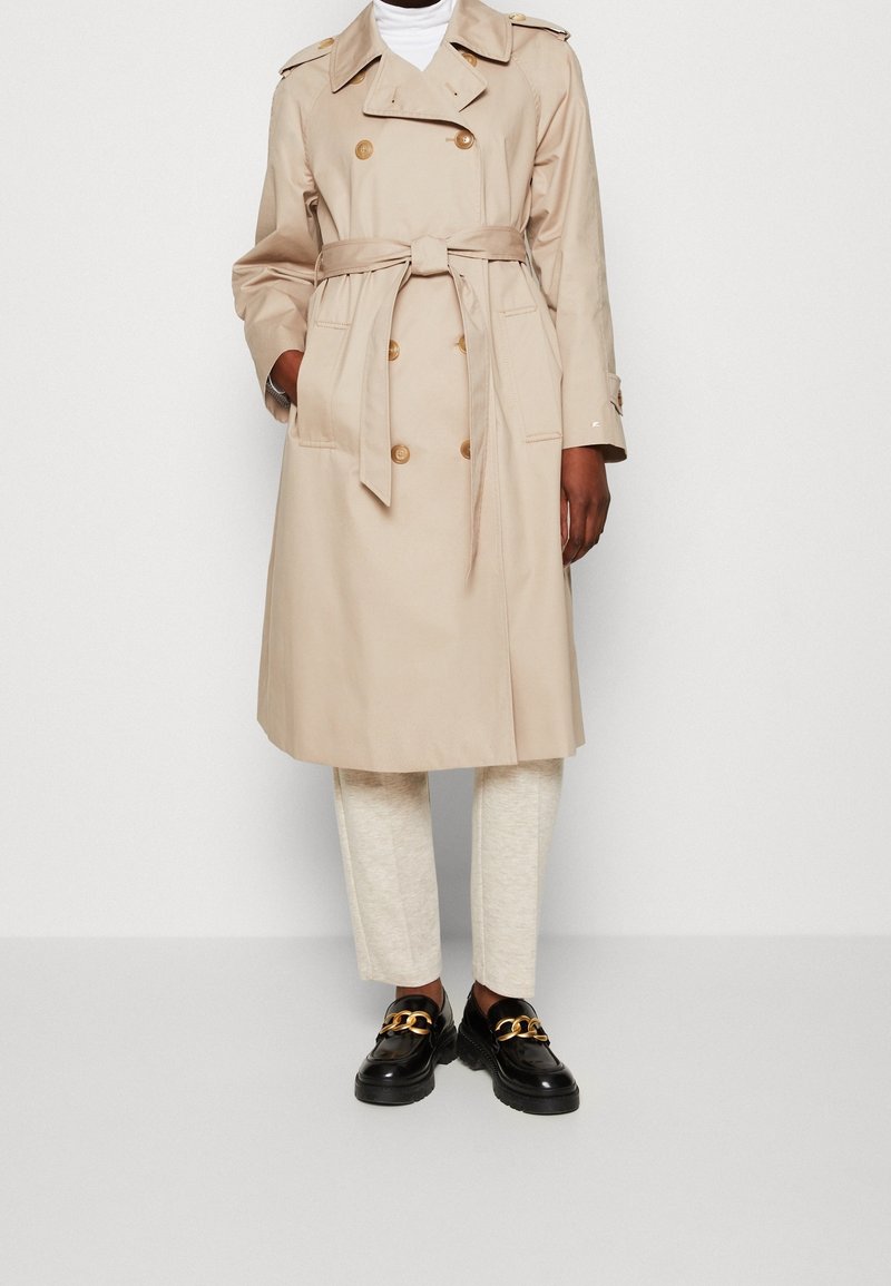 Personne portant un trench-coat beige à double boutonnage avec une ceinture à la taille, sur un pantalon crème et des mocassins noirs avec un détail de chaîne dorée.