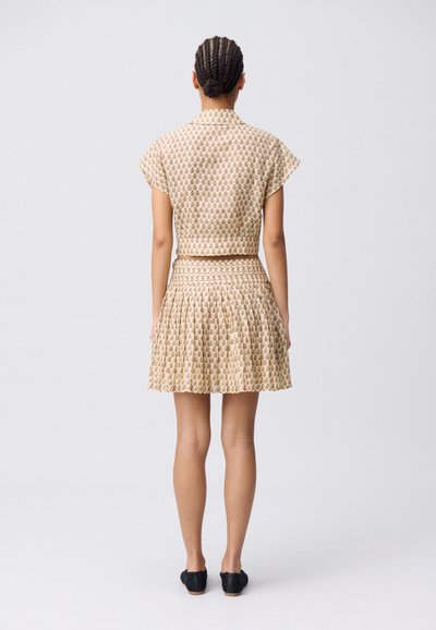 Completo composto da blusa corta a maniche corte e gonna plissettata in un tenue color nocciola con motivo floreale marrone; tessuto liscio con vestibilità comoda. Calzature nere.