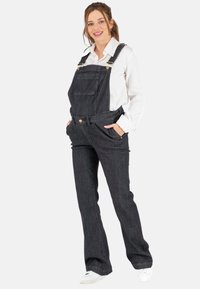 Dunkelblaue Jeanslatzhosen mit verstellbaren Trägern, einer geraden Vorderasche und schmal zulaufenden Beinen, kombiniert mit einem weißen Langarmshirt und weißen Turnschuhen.
