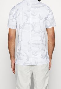 Muž v světle šedém tričku s krátkým rukávem se vzorem maskovacího efektu a béžových kalhotách, zobrazený zezadu na jednobarevném pozadí.