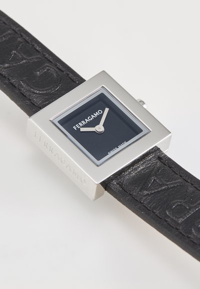 Négyzet alakú ezüst óra fekete bőrszíjjal, amelyen dombornyomott logó található. Sötét számlap ezüst mutatókkal és a "FERRAGAMO" logóval. Svájci gyártású.