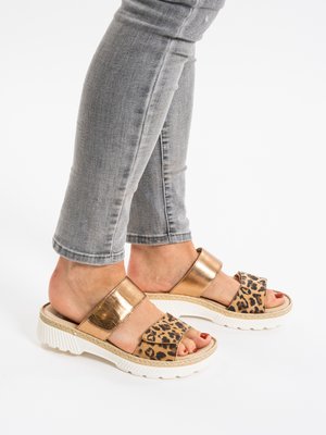 Pies con sandalias de punta abierta con tiras doradas y estampado de leopardo, combinadas con vaqueros grises remangados sobre un fondo blanco.