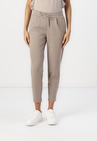 Beige tapered bukser lavet af blødt stof med snøre i taljen. Pletser foran og afsluttet med ankel-længde kanter. Parret med hvide sneakers.