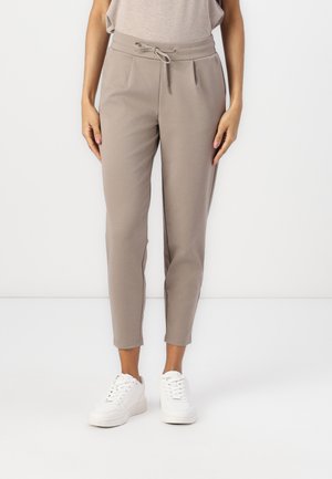 Beige tapered bukser lavet af blødt stof med snøre i taljen. Pletser foran og afsluttet med ankel-længde kanter. Parret med hvide sneakers.