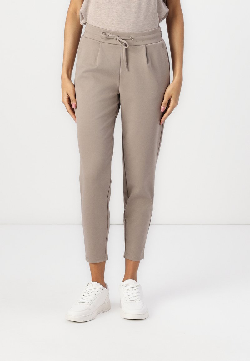 Beige tapered bukser lavet af blødt stof med snøre i taljen. Pletser foran og afsluttet med ankel-længde kanter. Parret med hvide sneakers.