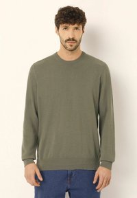 Pulover verde tricotat cu guler rotund, mâneci lungi și manșete și tiv cu margini ribbate. Textură moale cu o croială lejeră. Purtați peste blugi bleu.