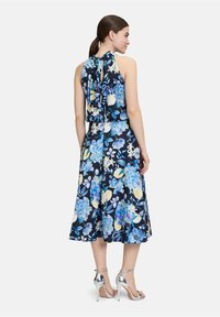 Blumenkleid in Midi-Länge mit dunklem Marineblau als Hintergrund, blauen und gelben Blumen, Halterneck und einem fließenden Rock. Silberne Sandalen mit Riemchen ergänzen das Outfit.