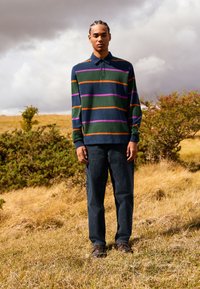 Námořnická polo košile s vodorovnými pruhy v zelené, oranžové a fialové barvě, v kombinaci s tmavými džínami a hnědými botami, postavená na poli.