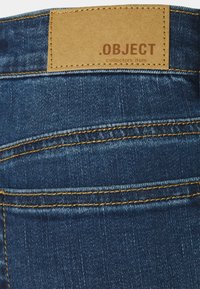 Los jeans de mezclilla azul oscuro presentan costuras doradas, una etiqueta de cuero con la inscripción ".OBJECT objeto de colección" y una superficie de textura suave.