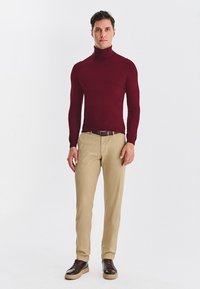 Bordowy sweter z golfem, dopasowany, w połączeniu z beżowymi spodniami i brązowymi trampkami. Gładka tekstura, minimalistyczny design, brak widocznych wzorów.