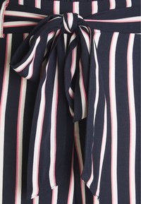 Námořnická a bílá pruhovaná látka s růžovými akcenty; má zavazovací pas a jemnou texturu. Ukazuje detailní mašličku a vertikální pruhy.