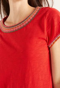 Femme portant un haut rouge à manches courtes orné d'un bord festonné coloré autour de l'encolure et des poignets, en rose, orange et vert.