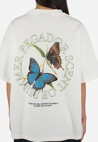 Camiseta blanca de gran tamaño con gráficos de mariposas coloridas, rodeada de texto verde. Cita en la parte inferior en negro, diseño simple.