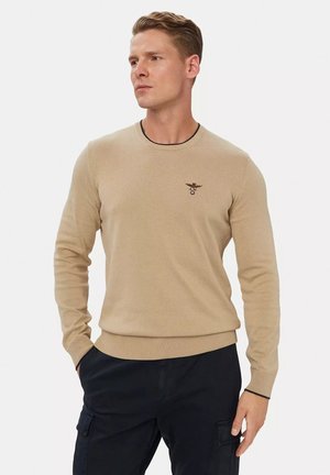 Uomo con capelli corti biondi che indossa un maglione beige con un piccolo logo sul petto e pantaloni neri con tasche sulle cosce.