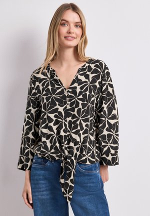 Jeune femme portant un chemisier à motif floral noir et beige avec un lien à l'avant, associé à un jean en denim bleu sur un fond uni.