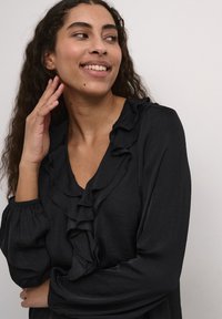 Black blouse with ruffled neckline and elastic sleeves. Lightweight fabric with a soft texture. Model poses with hand near face.