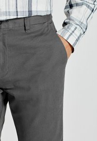 Pantalones grises texturizados de corte slim, con bolsillos laterales y una cinturilla suave, llevados con una camisa de cuadros de manga larga.