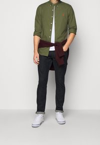 Mężczyzna w oliwkowej koszuli na guziki, białej podkoszulce, ciemnych dżinsach, białych trampkach i bordowym swetrze związanym w talii.