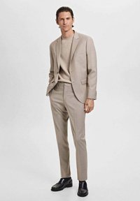 Light beige suit with tailored jacket and slim-fit trousers, paired with a cream sweater and black shoes. Smooth texture, minimal design details.