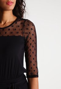 Zwarte top met stippenmotief op sheer mouwen en een effen lichaam. De mouwen zijn ellebooglang en hebben een gladde, lichte textuur.