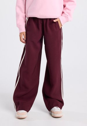 Person, der weinrote Hose mit weitem Bein und weißen Seitenstreifen, rosa Sweatshirt und weiße sowie rosa Turnschuhe trägt, steht mit einer Hand in der Tasche.