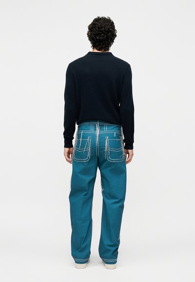 Άνδρας με σγουρά μαλλιά φοράει μαύρο πουκάμισο μακρυμάνικο και τιρκουάζ παντελόνι με έντονες λευκές ραφές, στέκεται γυρισμένος προς τα πίσω σε λευκό φόντο.