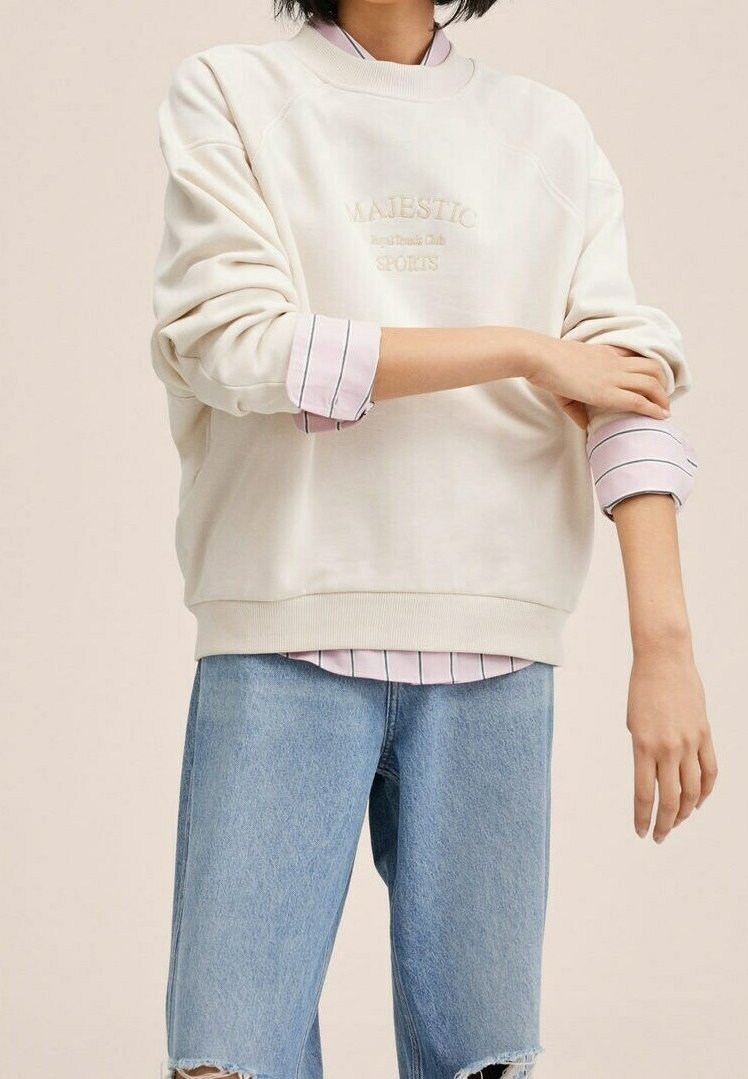 Cremerfarbenes Sweatshirt mit geprägtem Schriftzug "MAJESTIC". Kombiniert mit einem hellrosa gestreiften Hemd, das an den Manschetten hervorblitzt, und hellblauen, verwaschenen Shorts.