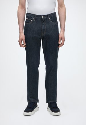 Mand iført mørkeblå jeans med lige ben og sorte og hvide sneakers, stående mod en ensfarvet hvid baggrund.