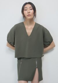 Top verde a maniche corte con scollo a V e maniche arricciate, abbinato a una gonna coordinata con zip frontali e uno spacco pronunciato.