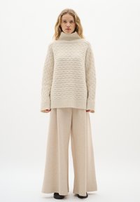 Cremefarbener Strickpullover mit hohem Kragen und strukturiertem Wellenmuster, kombiniert mit lockeren, beigen, weit geschnittenen Hosen und schwarzem Schuhwerk.