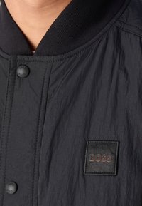 Gros plan d'une personne portant une veste noire avec des boutons-pression mats et un patch carré marqué BOSS sur la poitrine.