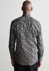 Pitkähihainen paita, jossa tumma sininen pohja ja monimutkaiset paisley-kuviot kullassa, beigeissä ja valkoisissa sävyissä. Silkkinen kangas, jossa istuva leikkaus.