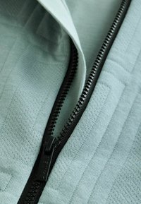 Hellgrüner Stoff mit einem strukturierten Muster, ausgestattet mit einem schwarzen Reißverschluss und Zipper. Der Stoff zeigt feine Webdetails und eine glatte Oberfläche.