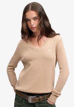 Jonge vrouw met lang golvend bruin haar, een beige V-hals trui en groene broek met een bruine riem, handen in de zakken.