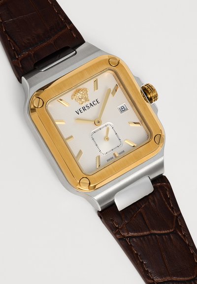 Montre-bracelet carrée Versace avec lunette dorée, cadran blanc, index dorés, affichage de la date et bracelet en cuir brun foncé à texture crocodile.