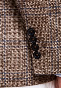 Manche de veste en tweed à carreaux marron et bleu avec quatre boutons noirs et un aperçu d’un poignet de chemise bleu clair.