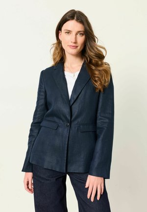 Femme aux longs cheveux ondulés portant un blazer bleu foncé par-dessus un haut blanc, debout devant un fond clair uni.