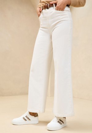 Person wearing high-waisted white wide-leg pants, brown belt, beige top, and white sneakers with leopard print accents.