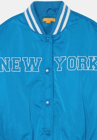 Blå varsityjacka med vit randig krage och tryckknappar, "NEW YORK" broderat över bröstet i vit omgiven text.