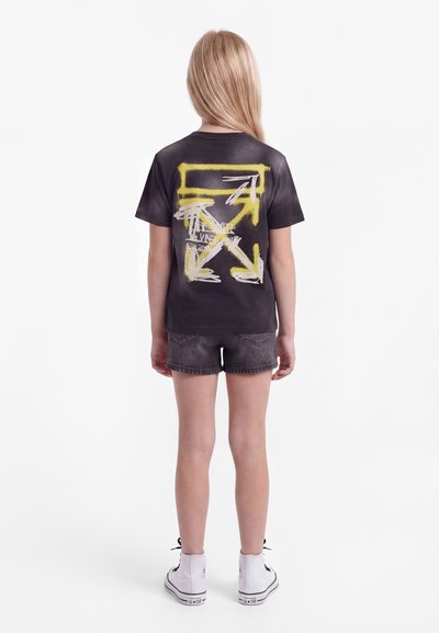 Kind mit langen blonden Haaren steht mit dem Rücken zur Kamera, trägt ein schwarzes T-Shirt mit gelbem und weißem Pfeildesign, dunkelblaue Shorts und weiße Turnschuhe.