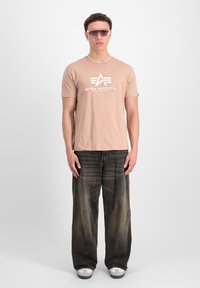 Beige kortærmet t-shirt med et hvidt logo, parret med løse, falmede sorte jeans og grå sneakers. Solbriller og halskæde inkluderet.