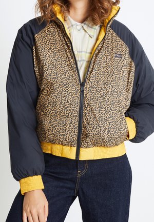 Femme portant une doudoune à imprimé léopard avec des manches noires, des poignets et un ourlet jaunes, par-dessus une chemise à carreaux claire et un jean foncé.