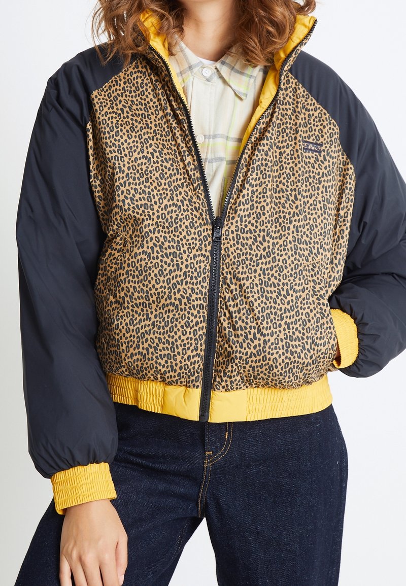 Femme portant une doudoune à imprimé léopard avec des manches noires, des poignets et un ourlet jaunes, par-dessus une chemise à carreaux claire et un jean foncé.