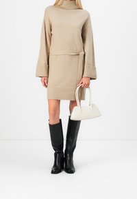 Robe en maille beige avec un col montant et une taille ceinturée, dotée de manches larges. Bottes noires montantes jusqu'au genou et sac à main crème.
