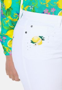 Jeans bianchi con limoni gialli ricamati e foglie verdi, arricchiti da piccoli strass. Il tessuto sembra liscio e elastico.